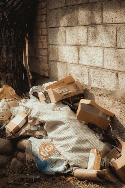 An outdoor scene showing a pile of assorted rubbish in a small designated waste collection area. The waste includes black plastic garbage bags, some torn open revealing mixed household refuse, along w
